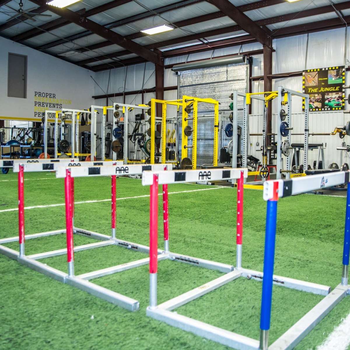 A training facility with agility hurdles positioned in front of a gym area equipped with weightlifting racks and exercise machines.