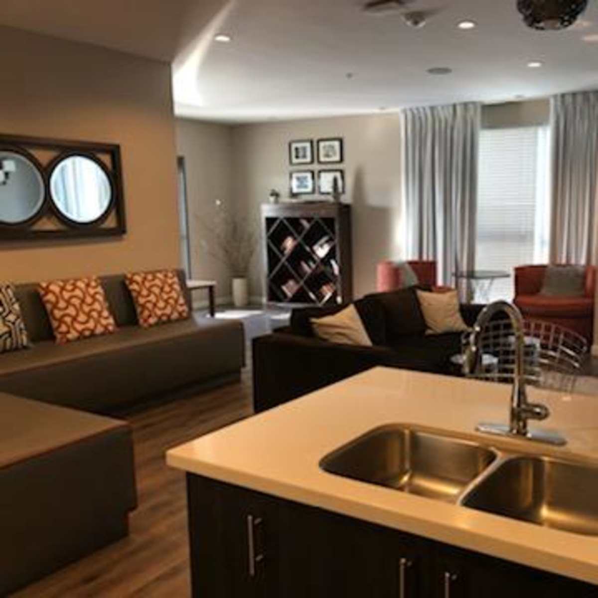 A modern living area with a kitchen island featuring a double sink, seating areas with couches and chairs, and large windows covered by curtains.