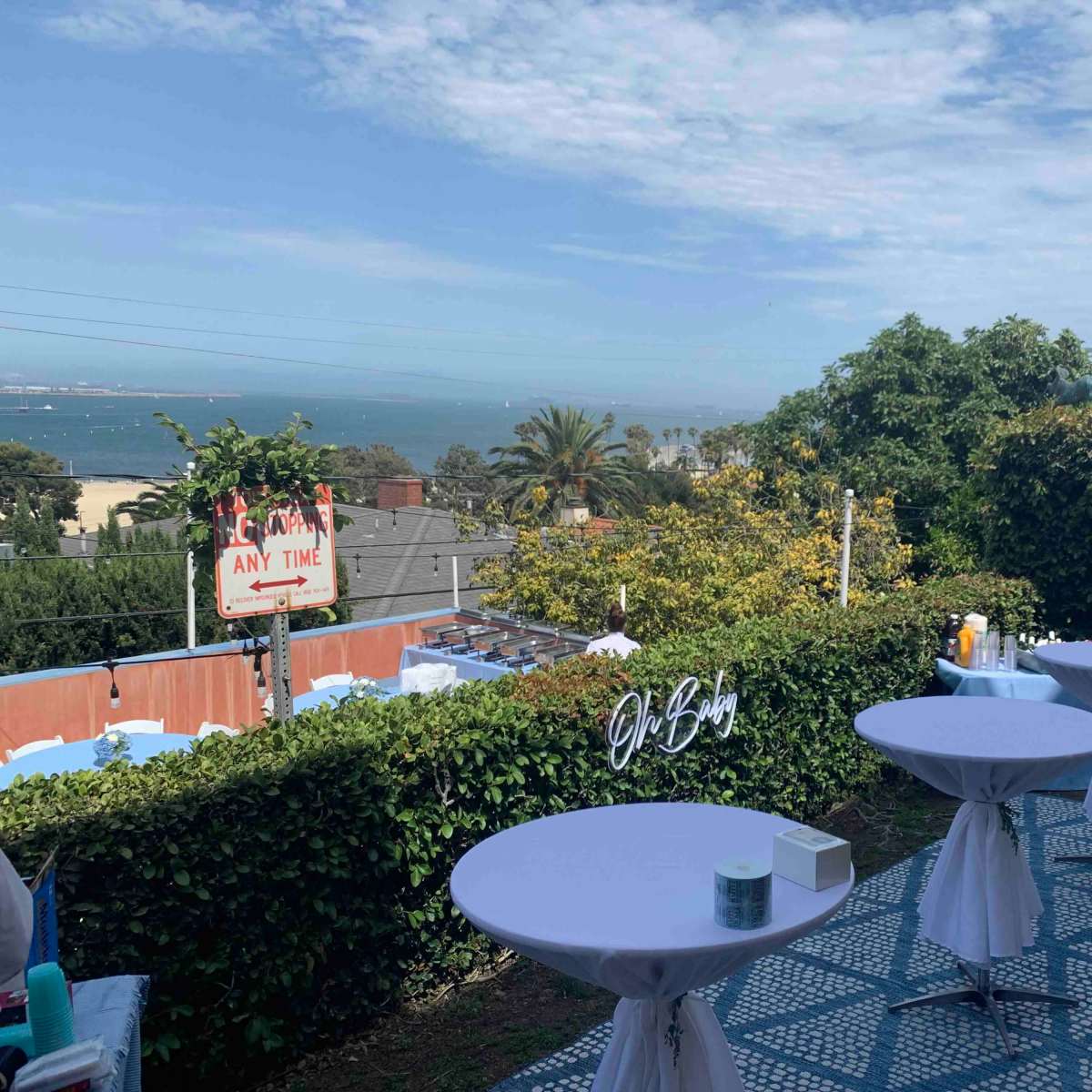 The scene shows a patio area set up for an outdoor event, featuring several round tables with white tablecloths, overlooking a view of the ocean and greenery.