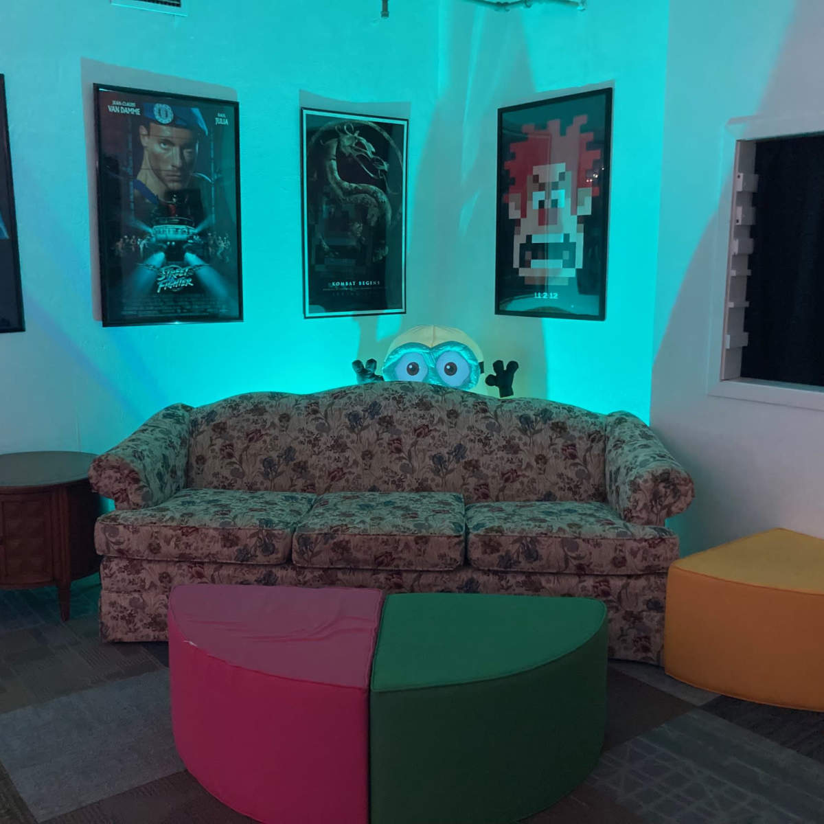 A room with a floral-patterned couch, colorful segmented seating, and movie posters on the walls under teal lighting.