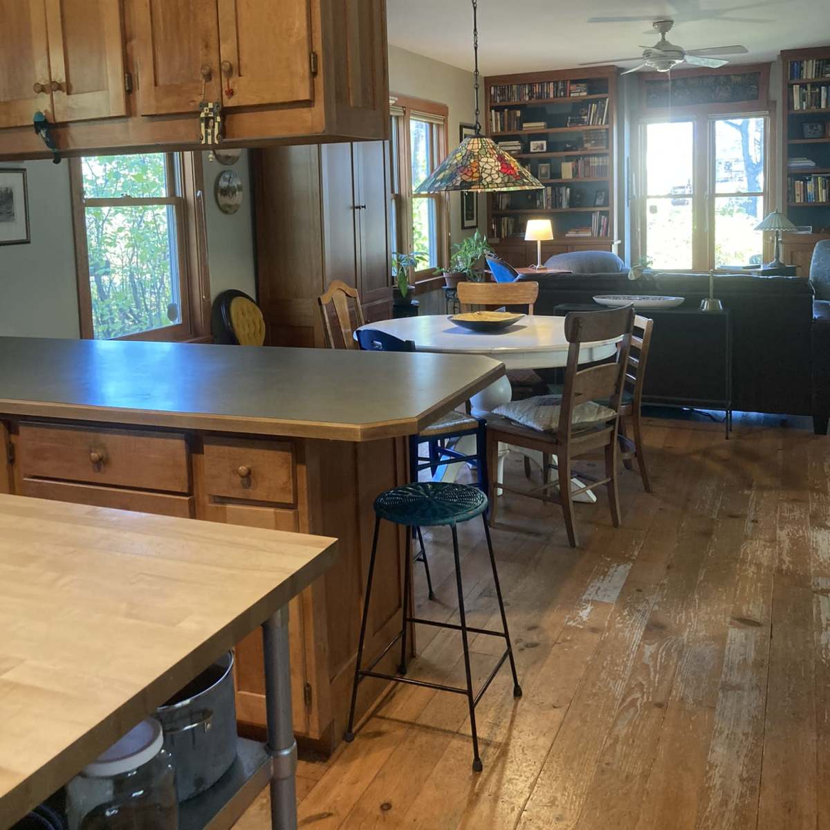 A wooden kitchen with a central island, a dining table surrounded by chairs, and a living area visible through large windows.