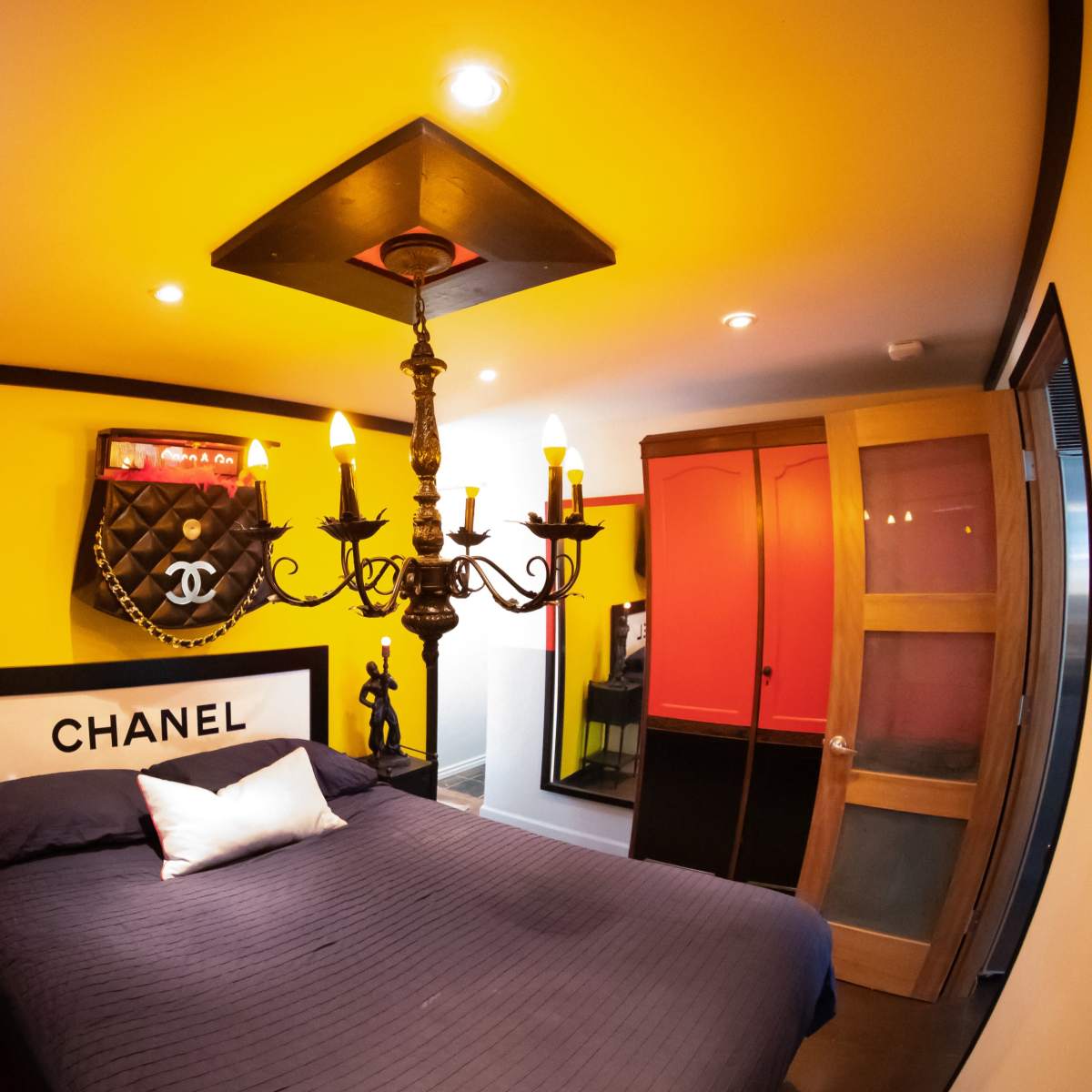 A brightly colored bedroom featuring a black and white Chanel-themed bed, a chandelier, and a closet with red doors.