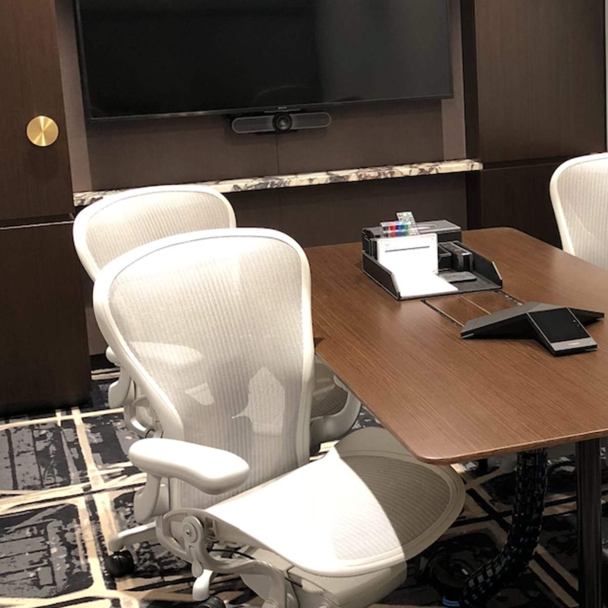 A modern conference room with a wooden table, four white ergonomic chairs, and a wall-mounted television.