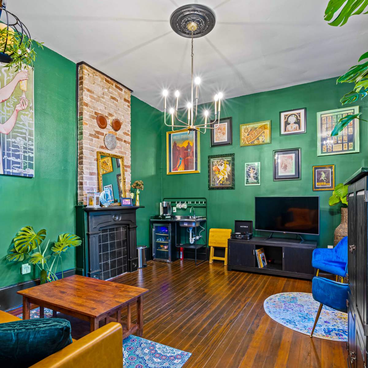 A living room with green walls, a brick accent wall, a chandelier, various framed artworks, a television, and a mix of furniture including a digital workstation and seating.