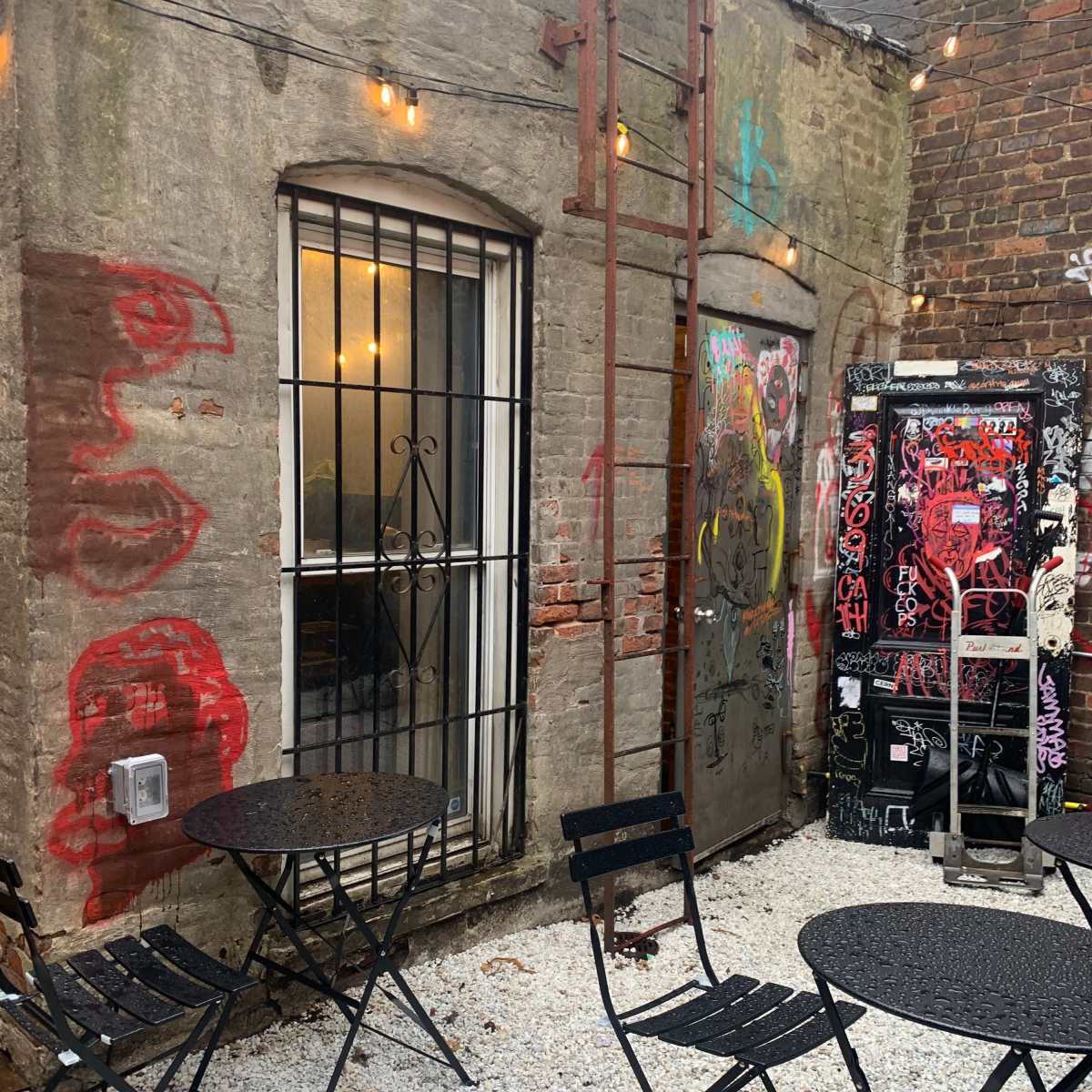A small outdoor seating area with black metal tables and chairs is set against a graffiti-covered wall and an adjacent building with a barred window.