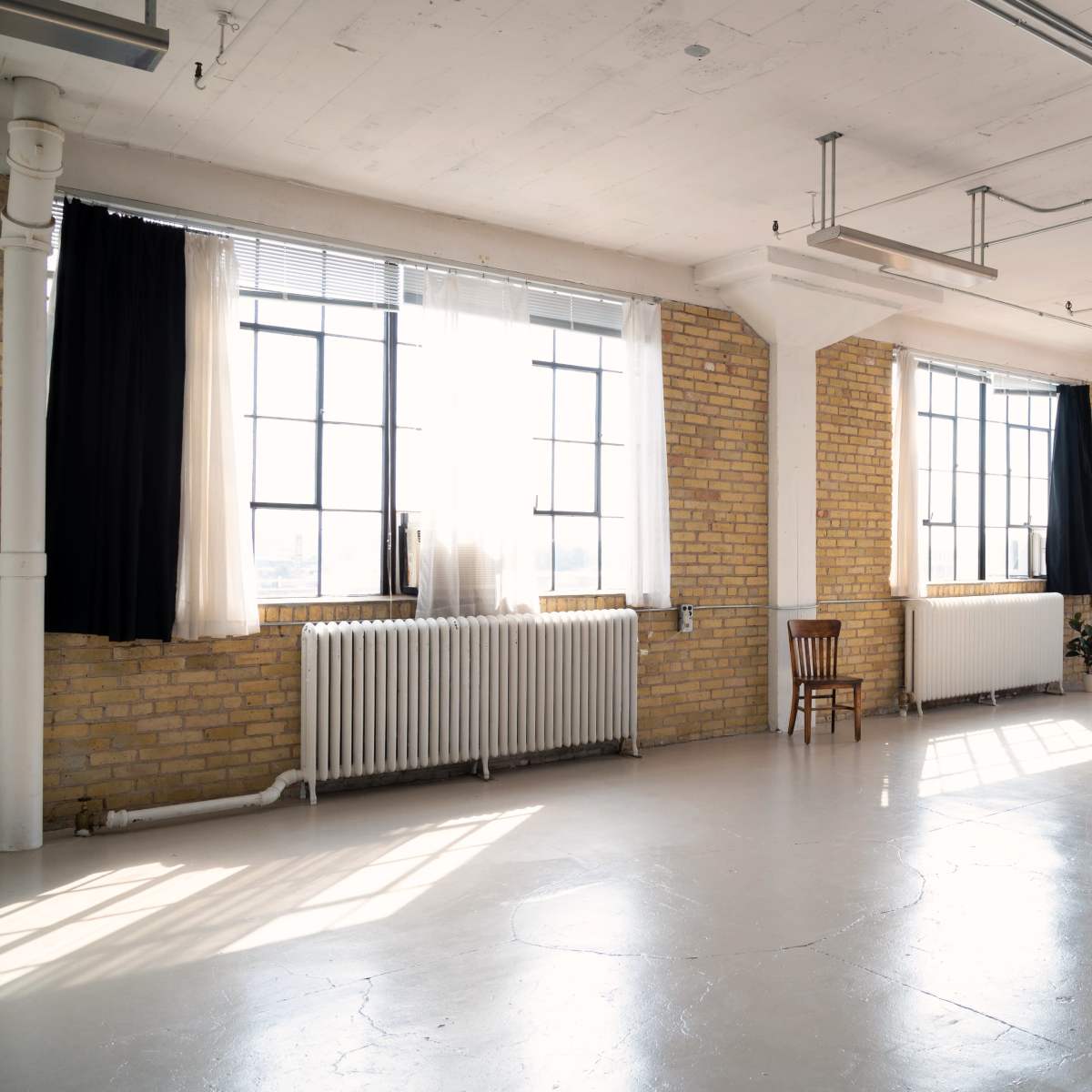 A spacious, empty room with large windows, exposed brick walls, a wooden chair, and a mirror.