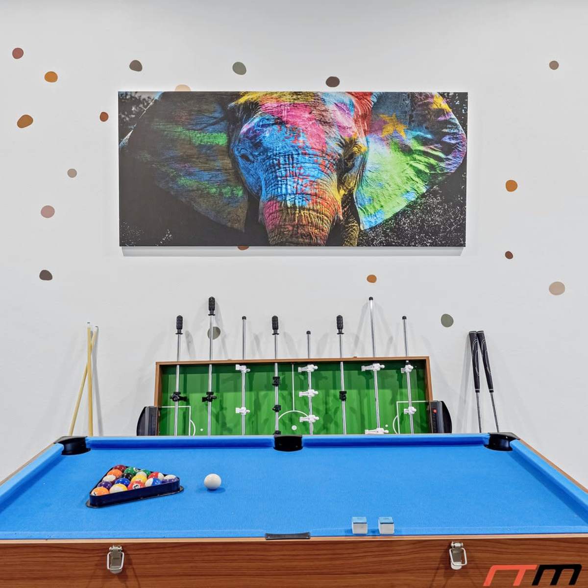A game room featuring a blue pool table, a foosball table, and a colorful elephant artwork on the wall.