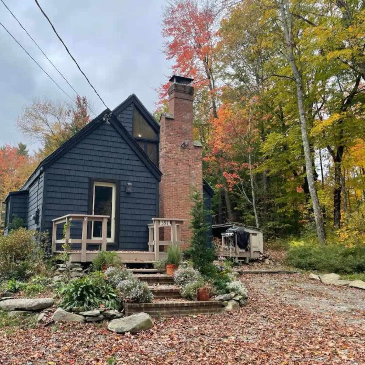 A two-story house with a brick chimney and dark siding is surrounded by trees with autumn foliage.