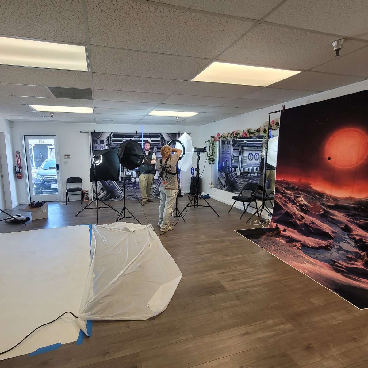 A photographer is setting up a lighting kit in a studio with a backdrop of a space scene and a blank white wall.