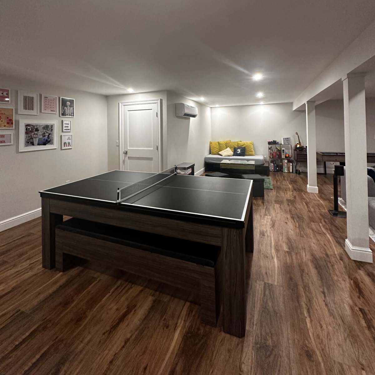A basement recreation room with a ping pong table in the center and a couch in the background.