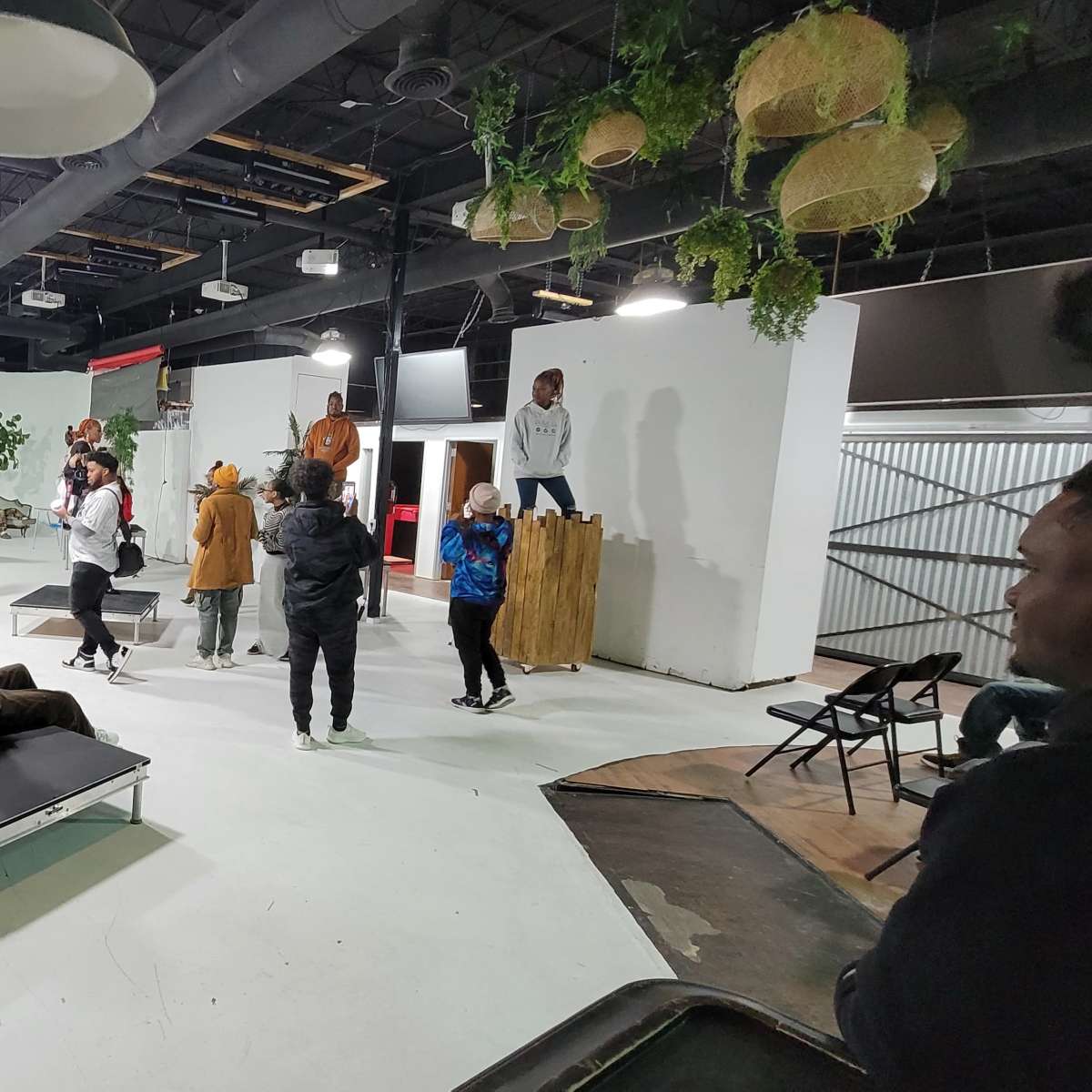 A group of people is engaged in various activities inside a spacious, indoor studio featuring greenery and wooden structures.