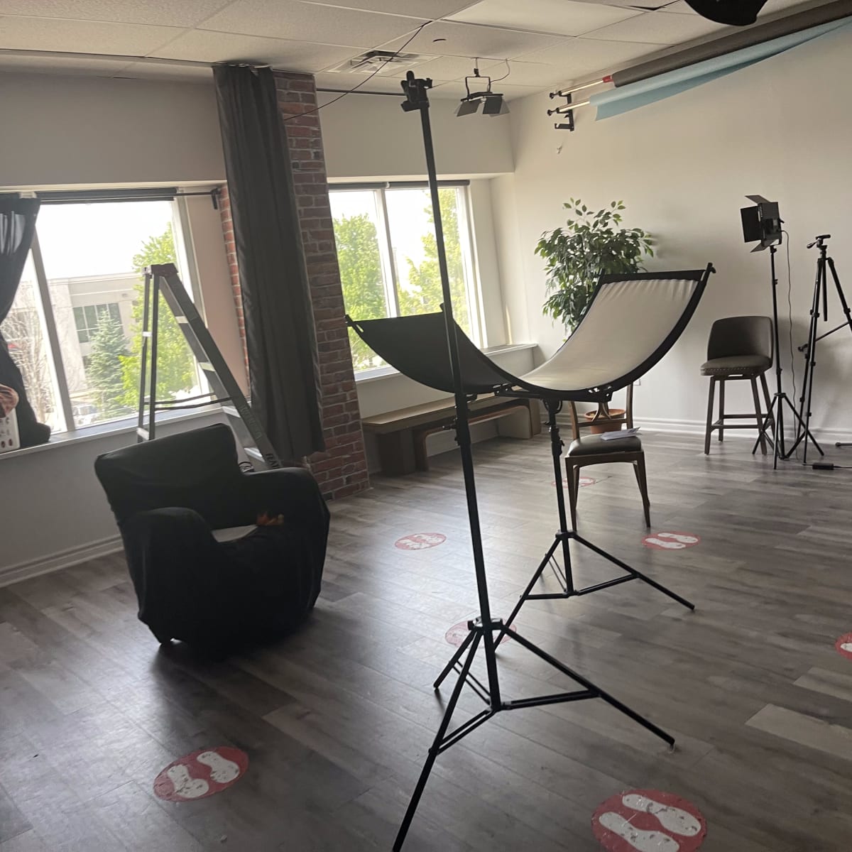 A photography studio featuring a black chair, a hammock stand, and several lighting equipment setups, with large windows allowing natural light.
