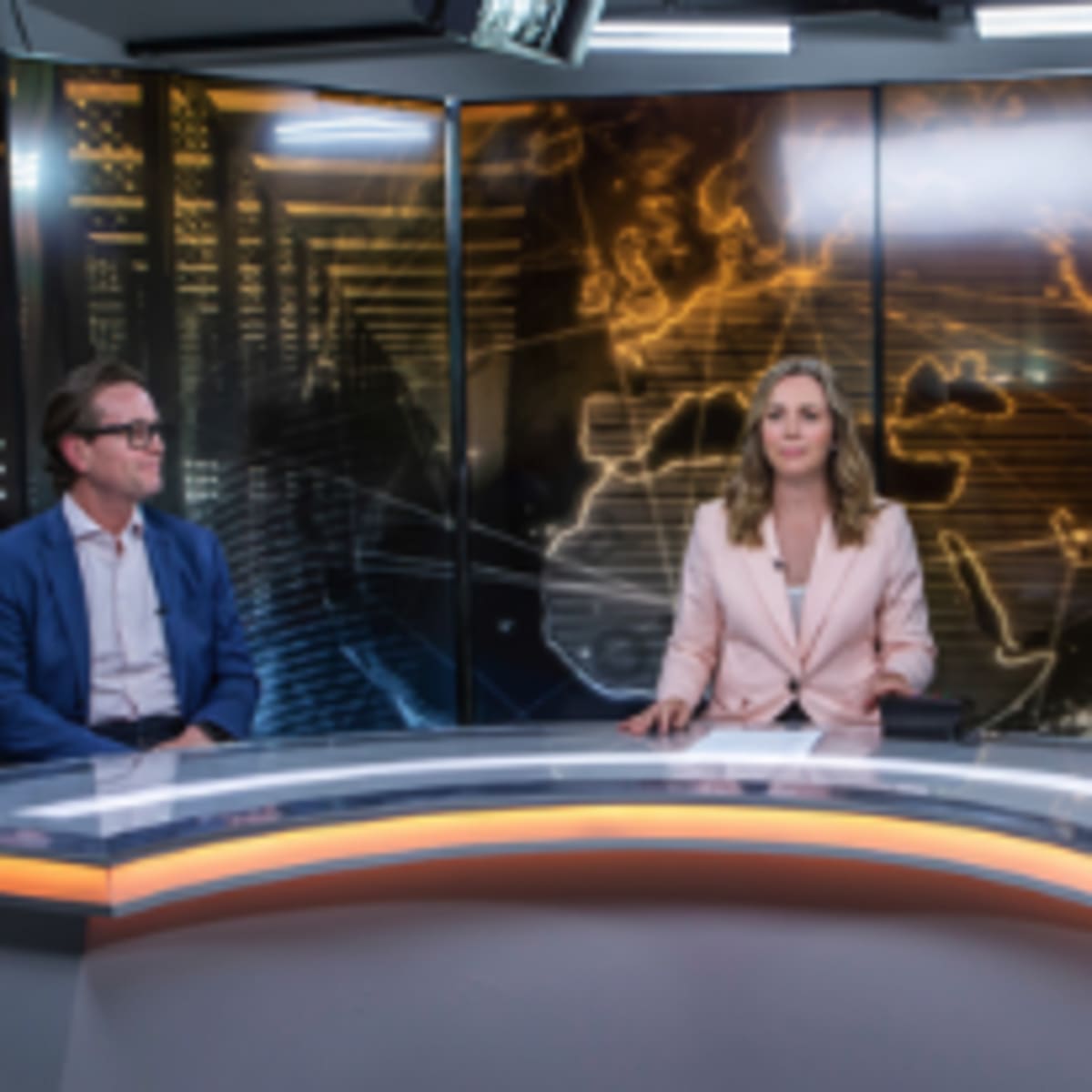 A news studio features three presenters seated at a curved table with a digital backdrop depicting a map and data graphics.