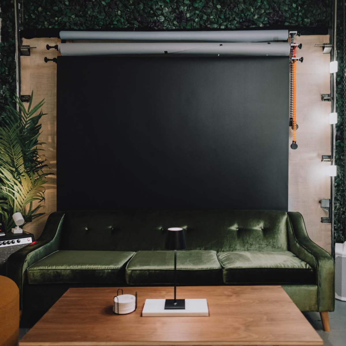A green velvet sofa facing a wooden table in a room with a black backdrop and plant decor.