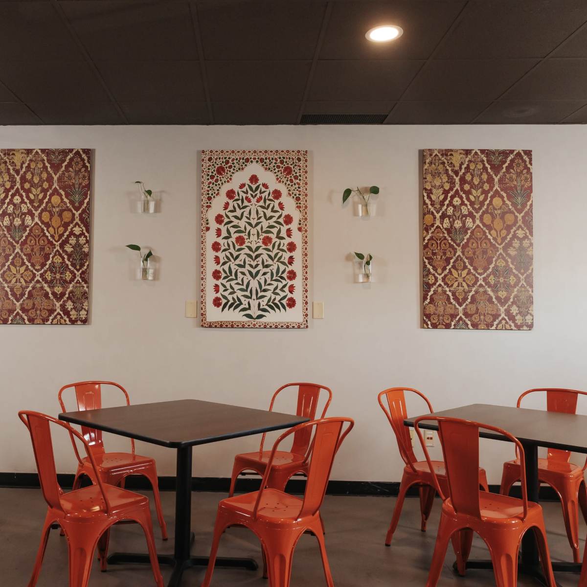 A modern café interior with orange chairs around black tables, featuring decorative wall art and small plants in hanging containers.