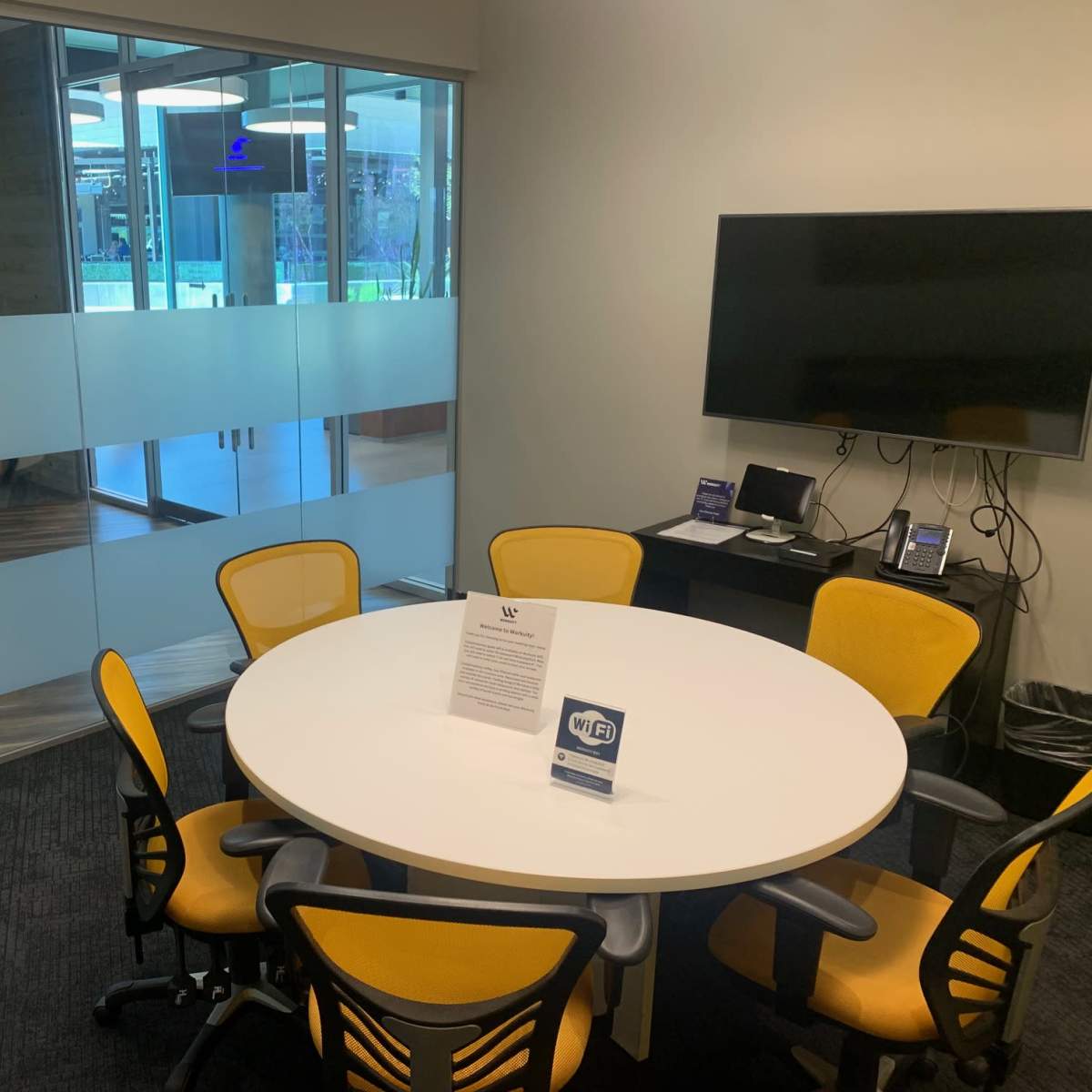 A round table with six yellow chairs is situated in a small meeting room featuring a wall-mounted television and glass partitions.