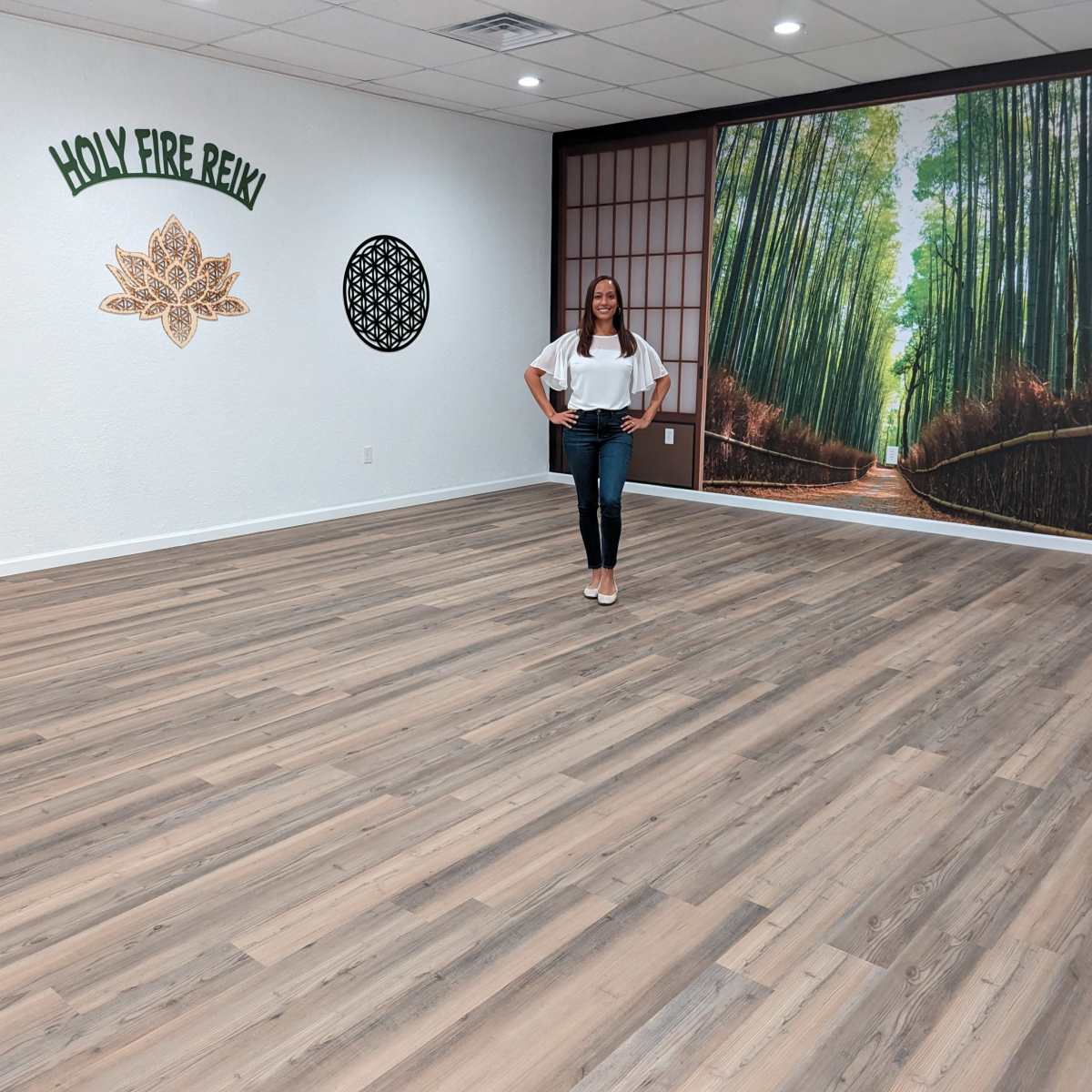 A woman stands on a wooden floor in a brightly lit yoga studio featuring a large bamboo mural and decorative wall art.