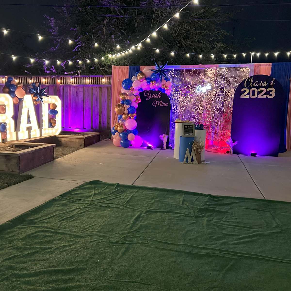 The scene features a decorated outdoor space for a graduation celebration, including illuminated letters spelling "GRAD," a backdrop with "Class of 2023," and colorful balloons.
