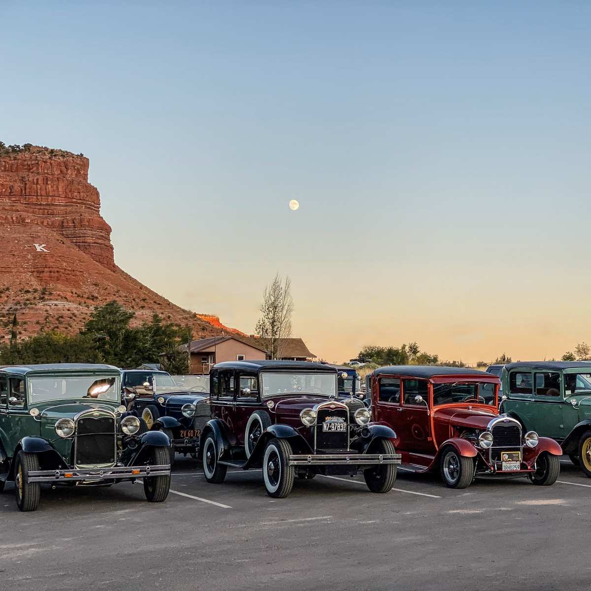 A line of vintage cars from the 1920s is parked in a lot with a rocky cliff and a full moon in the background.