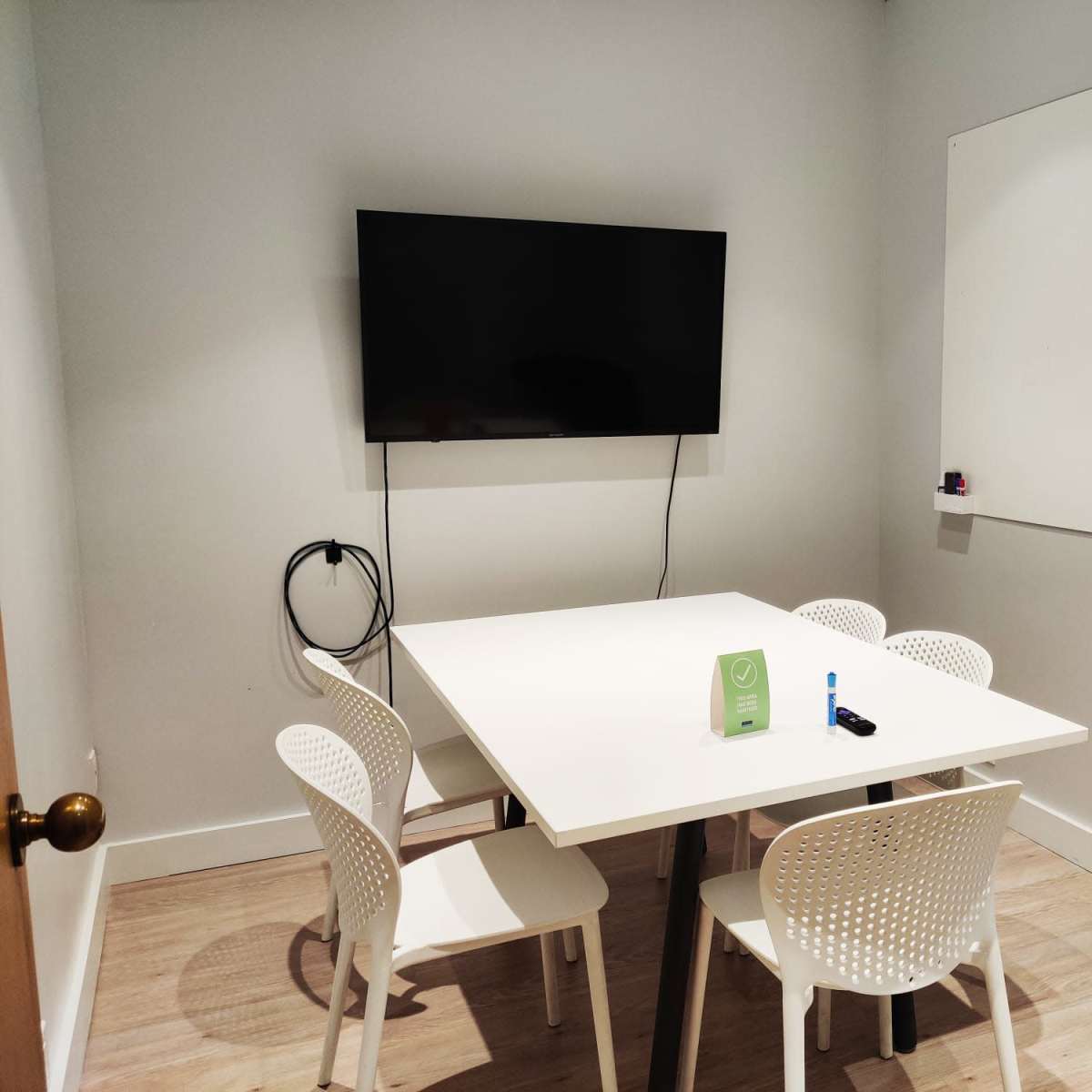 A small meeting room features a white table surrounded by four chairs, a television mounted on the wall, and a whiteboard.