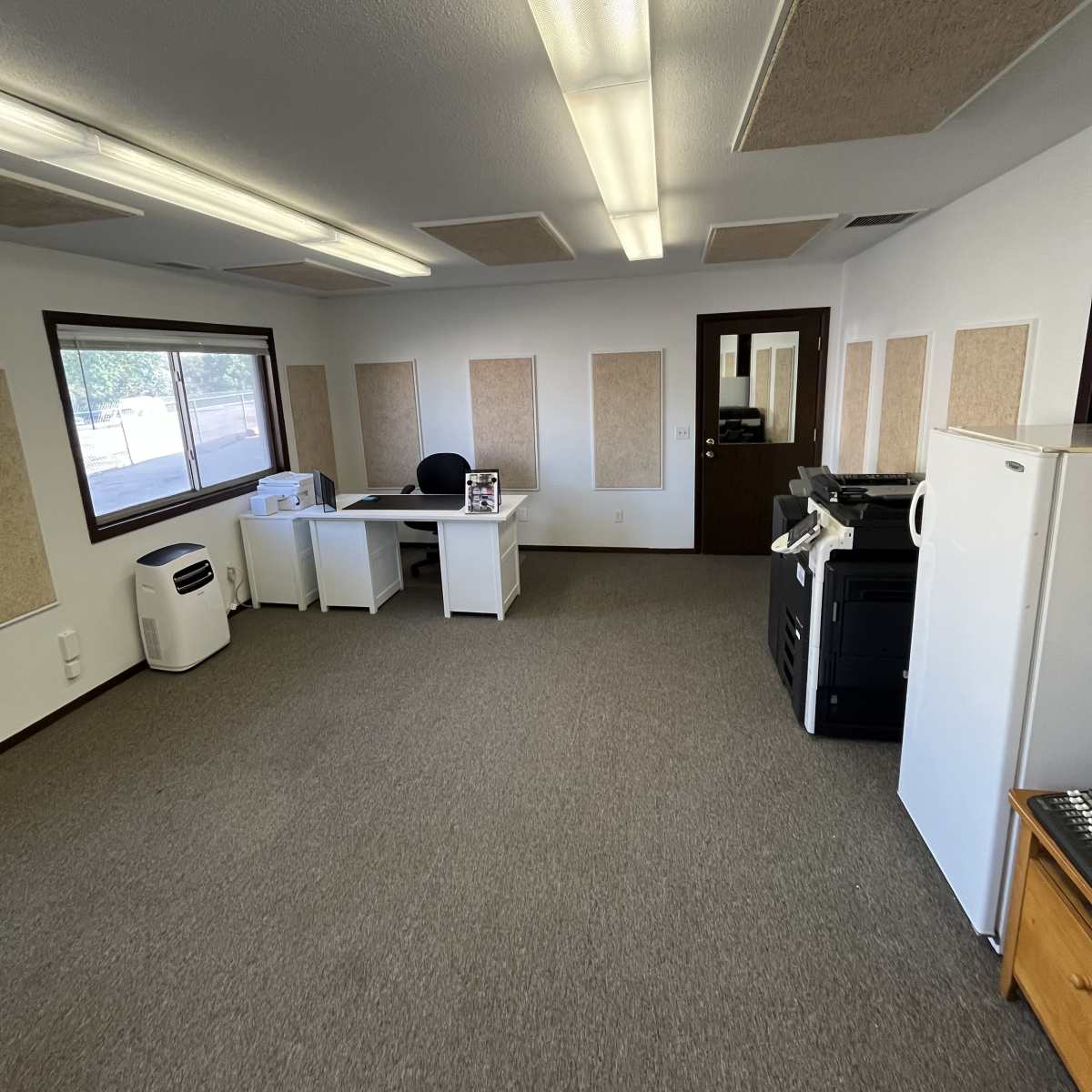 An empty office space features a white desk, office chair, refrigerator, and printer against the walls, with windows allowing natural light.