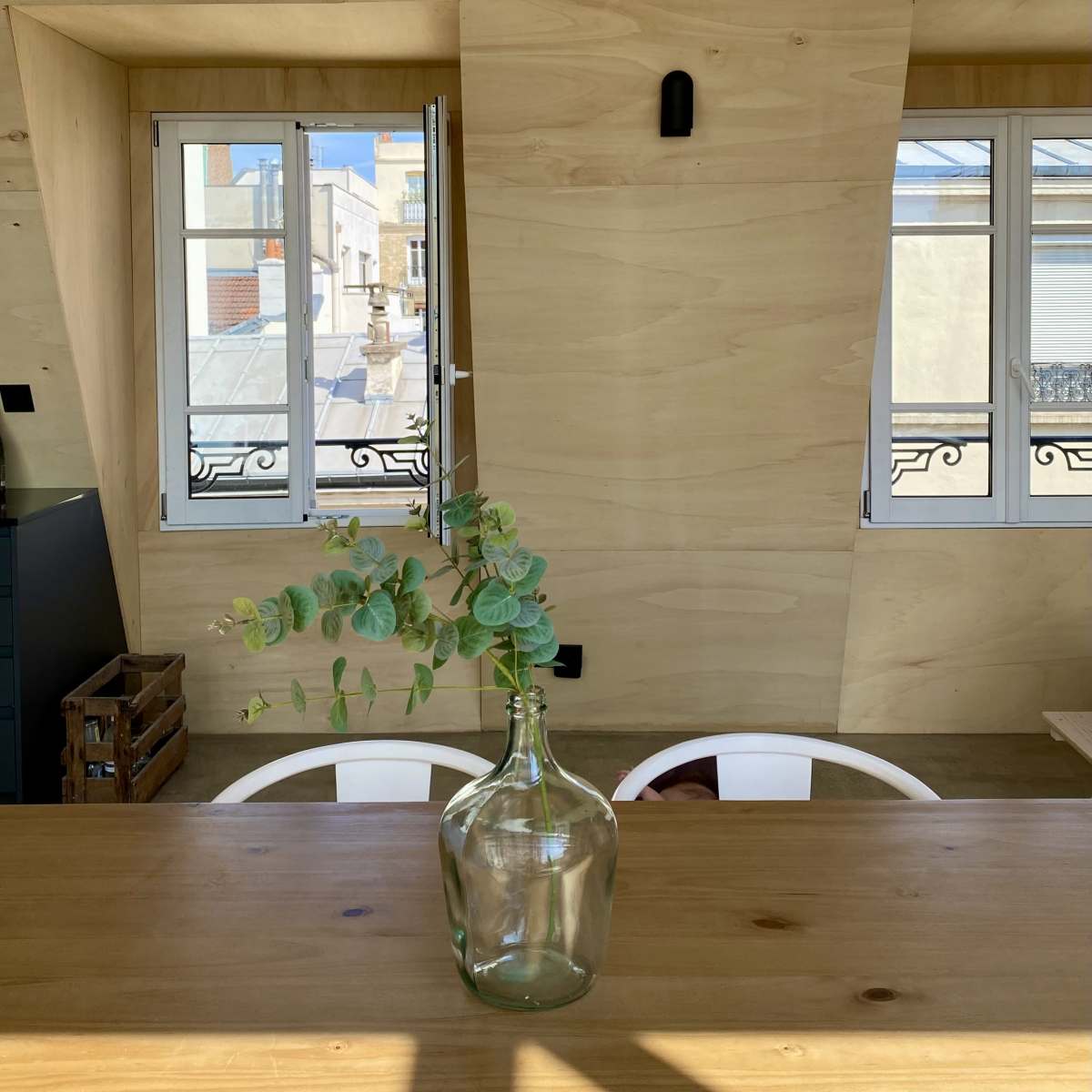 A minimalist wooden interior with a glass vase containing eucalyptus placed on a table, and two windows revealing a view of rooftops outside.