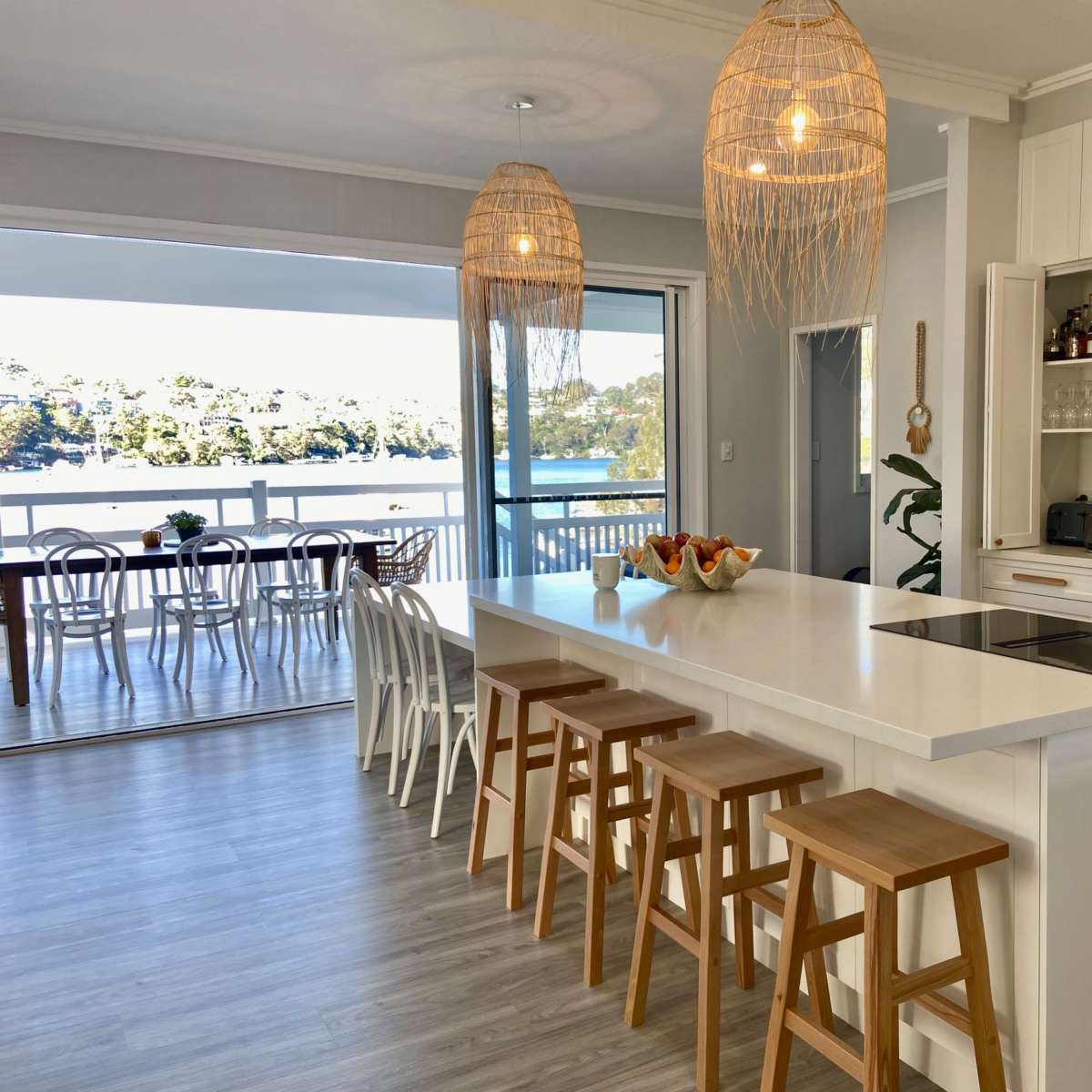 A modern kitchen features a central island with wooden stools and pendant lighting, overlooking a dining area and a scenic view of water and greenery.