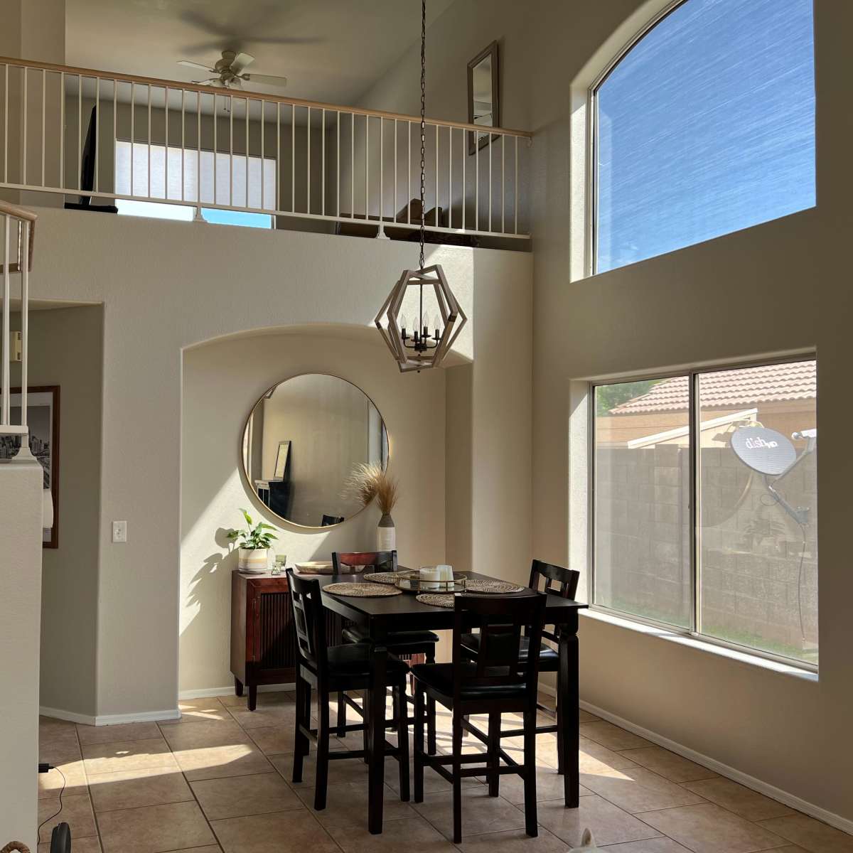 A spacious dining area with a round mirror and a black dining table set against a backdrop of large windows and a staircase.