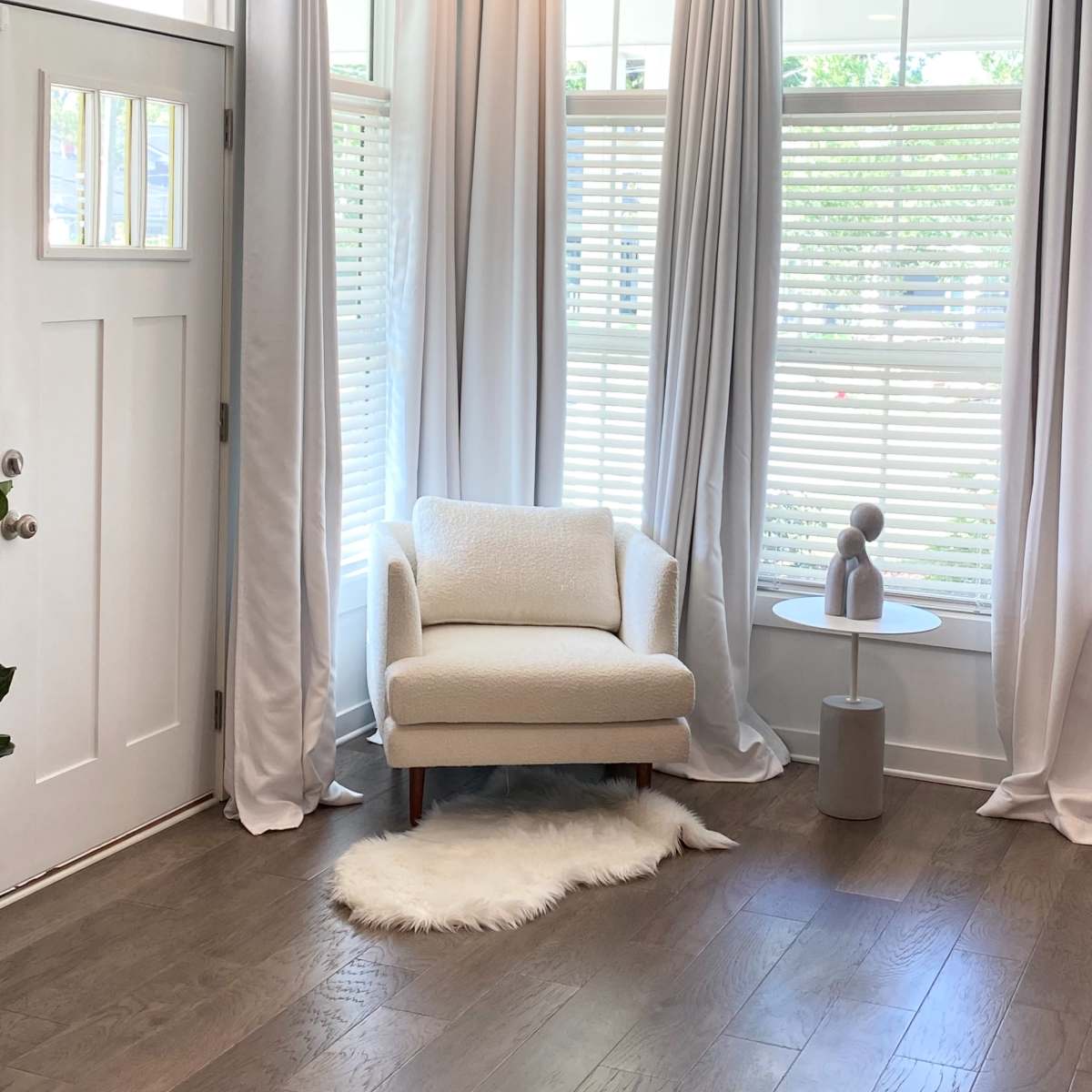 A bright corner of a room featuring a light-colored chair next to a small round table, with sheer white curtains framing the windows.