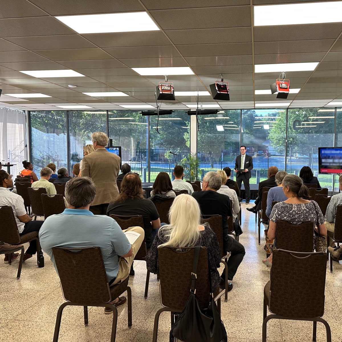 A speaker presents to an audience seated in a conference room with large windows overlooking greenery.