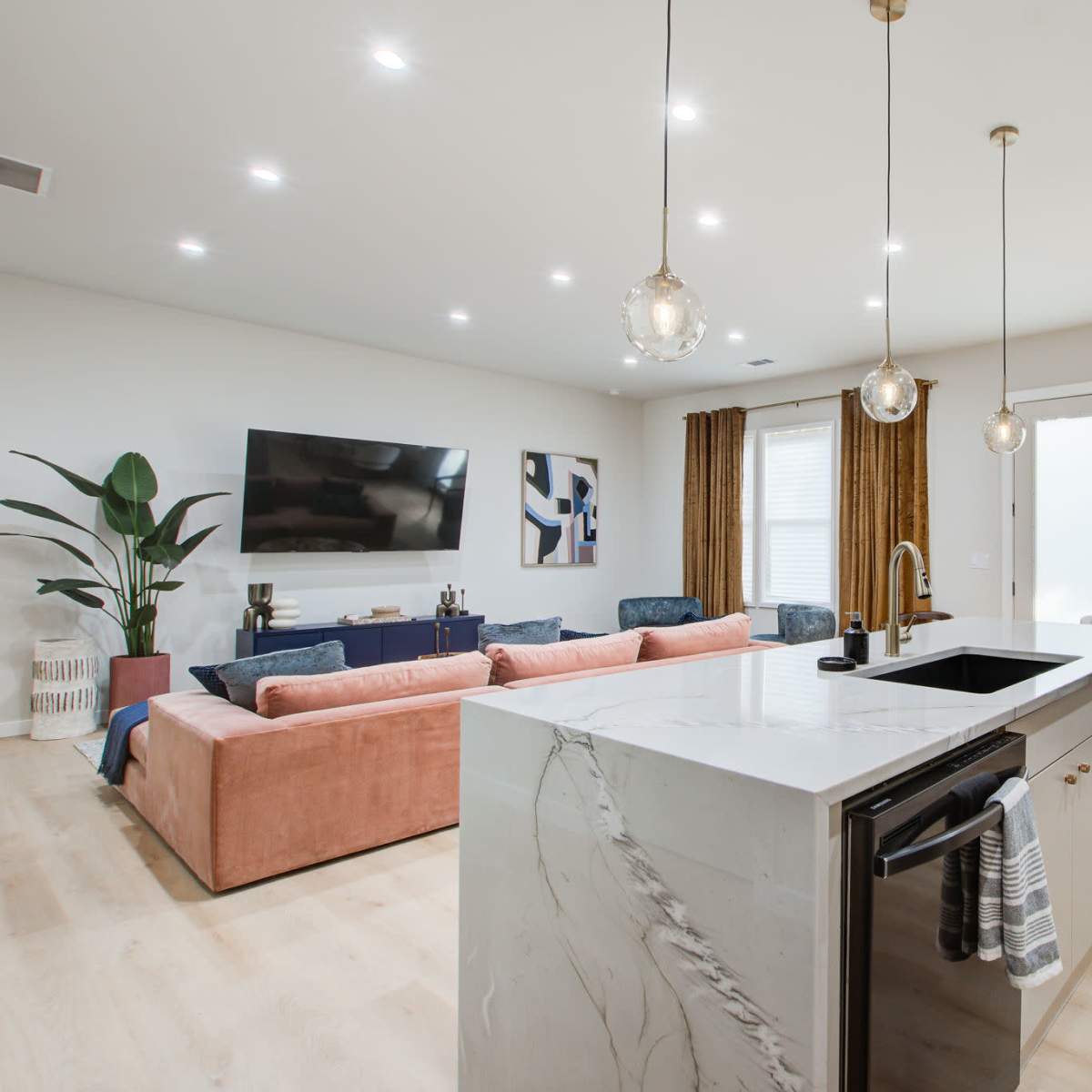 A modern living space featuring a kitchen island with pendant lights, a seating area with a sofa, and a dining table beside large windows.