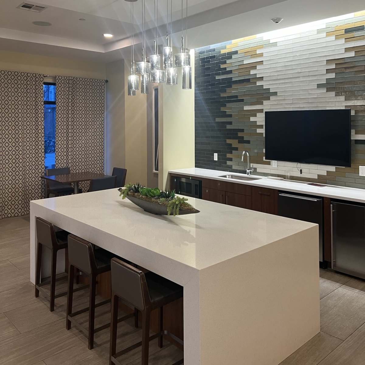 A modern kitchen and dining area featuring a large white countertop, a row of pendant lights overhead, and a wall with decorative tiles.