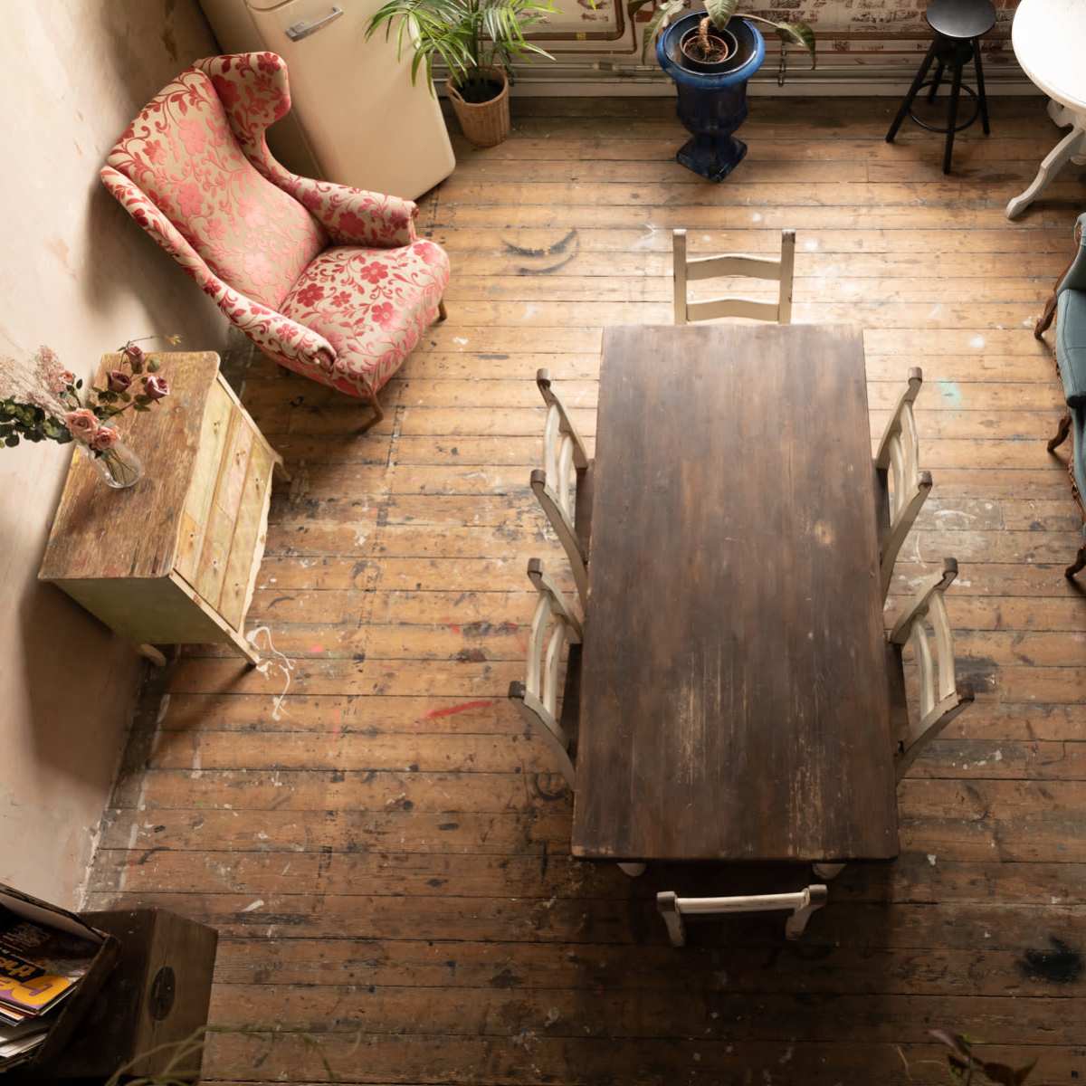 An overhead view of a rustic dining area featuring a wooden table surrounded by chairs, a floral armchair, and a green loveseat, with various plants and furniture against textured walls and a wooden floor.
