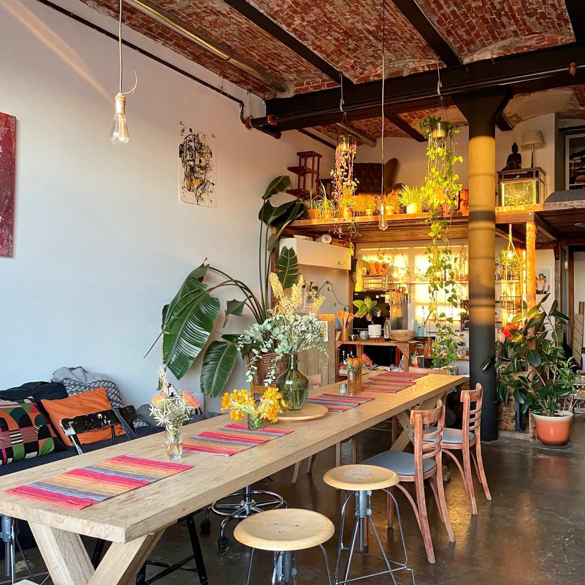A spacious loft features a long wooden dining table surrounded by chairs, with plants and artistic decorations visible in the background.