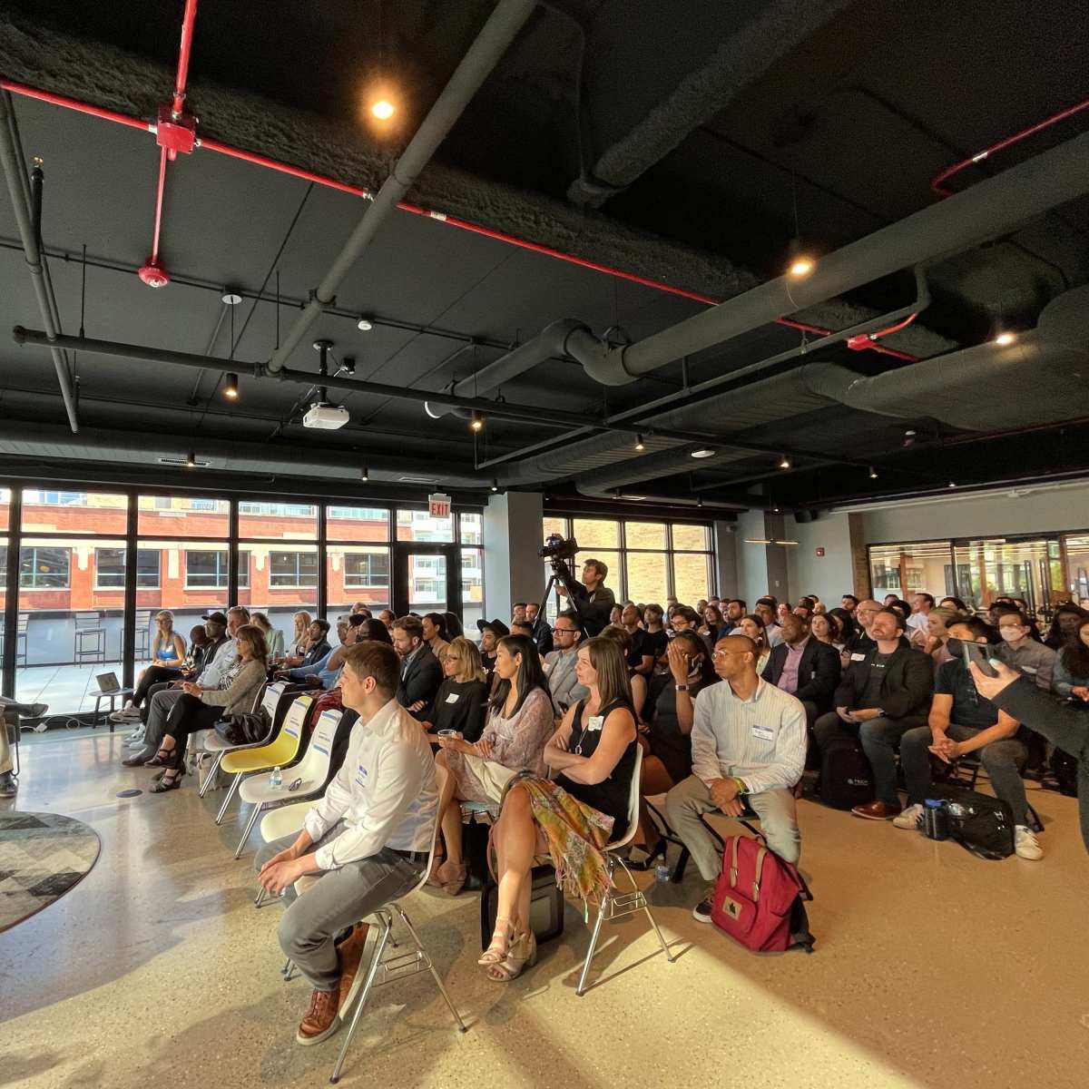 A diverse group of people sits in a modern office space, attentively watching a presentation on a screen at the front.