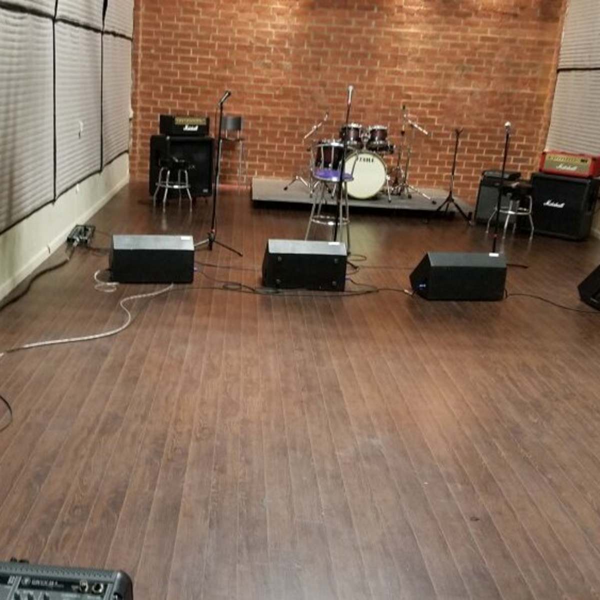 A music rehearsal space with a drum set on a raised stage, several amplifiers, and a row of empty chairs arranged in front.