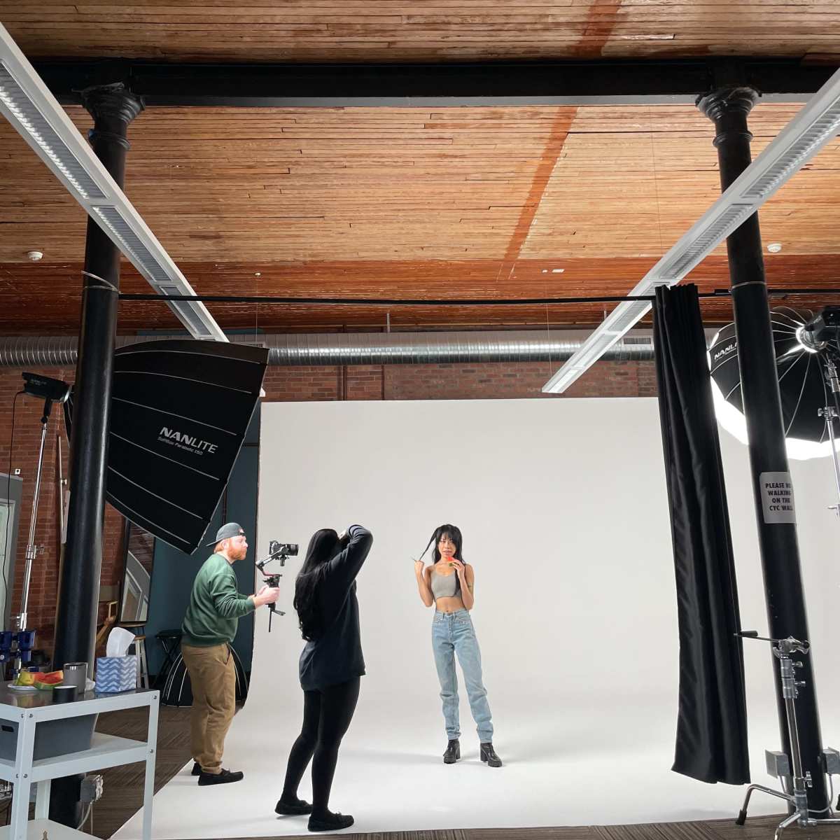 A photoshoot is taking place in a studio with a model posing in front of a white backdrop while a photographer and another crew member set up equipment.