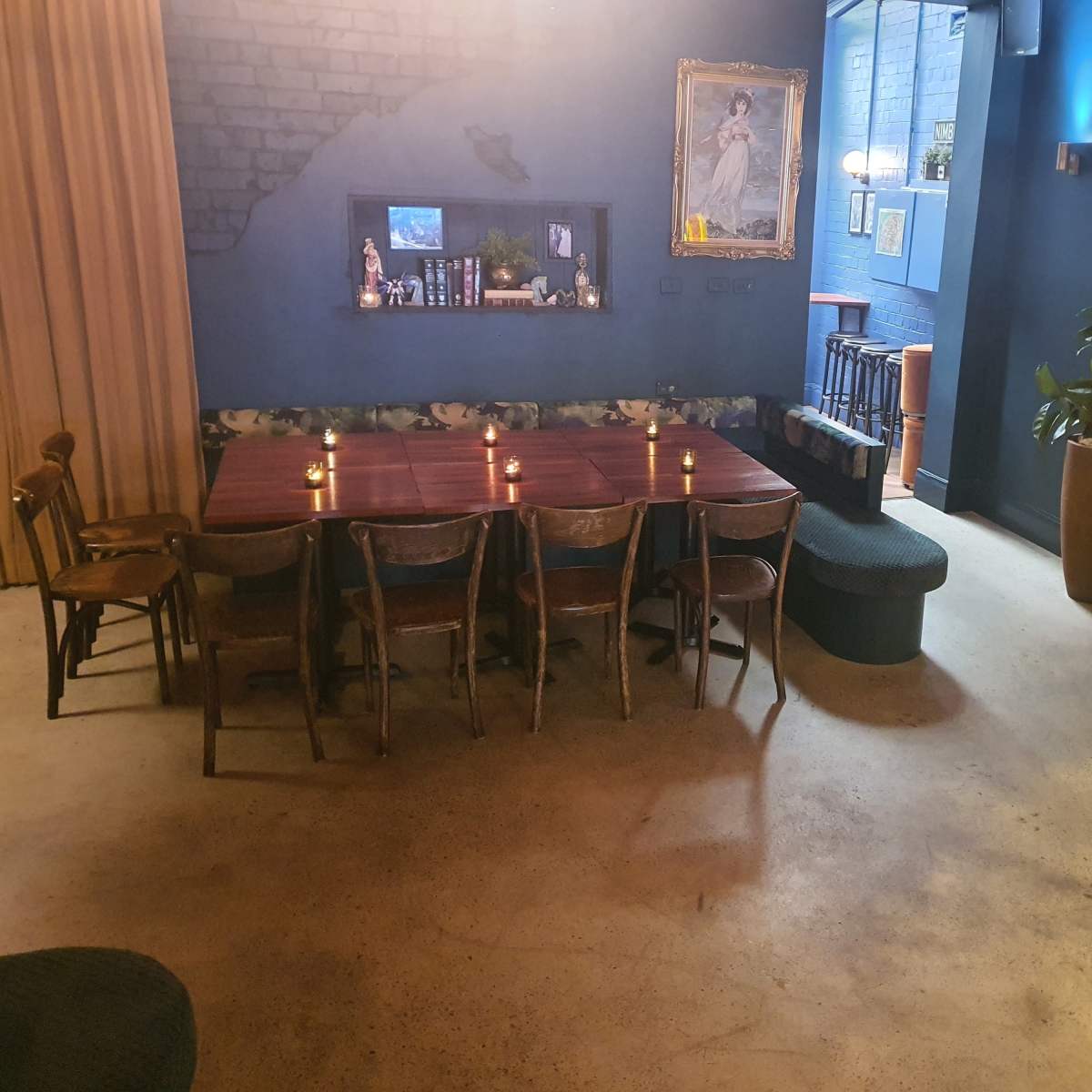 A dimly lit room with a long wooden table surrounded by chairs, a corner seating area, and a jukebox against a blue wall.