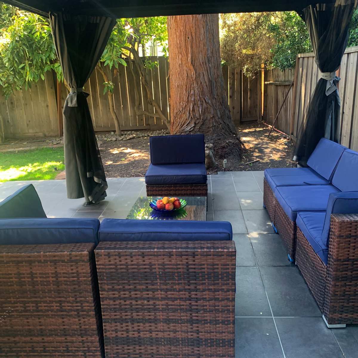 A shaded outdoor seating area with wicker furniture arranged around a glass-topped table, surrounded by greenery and wooden fencing.
