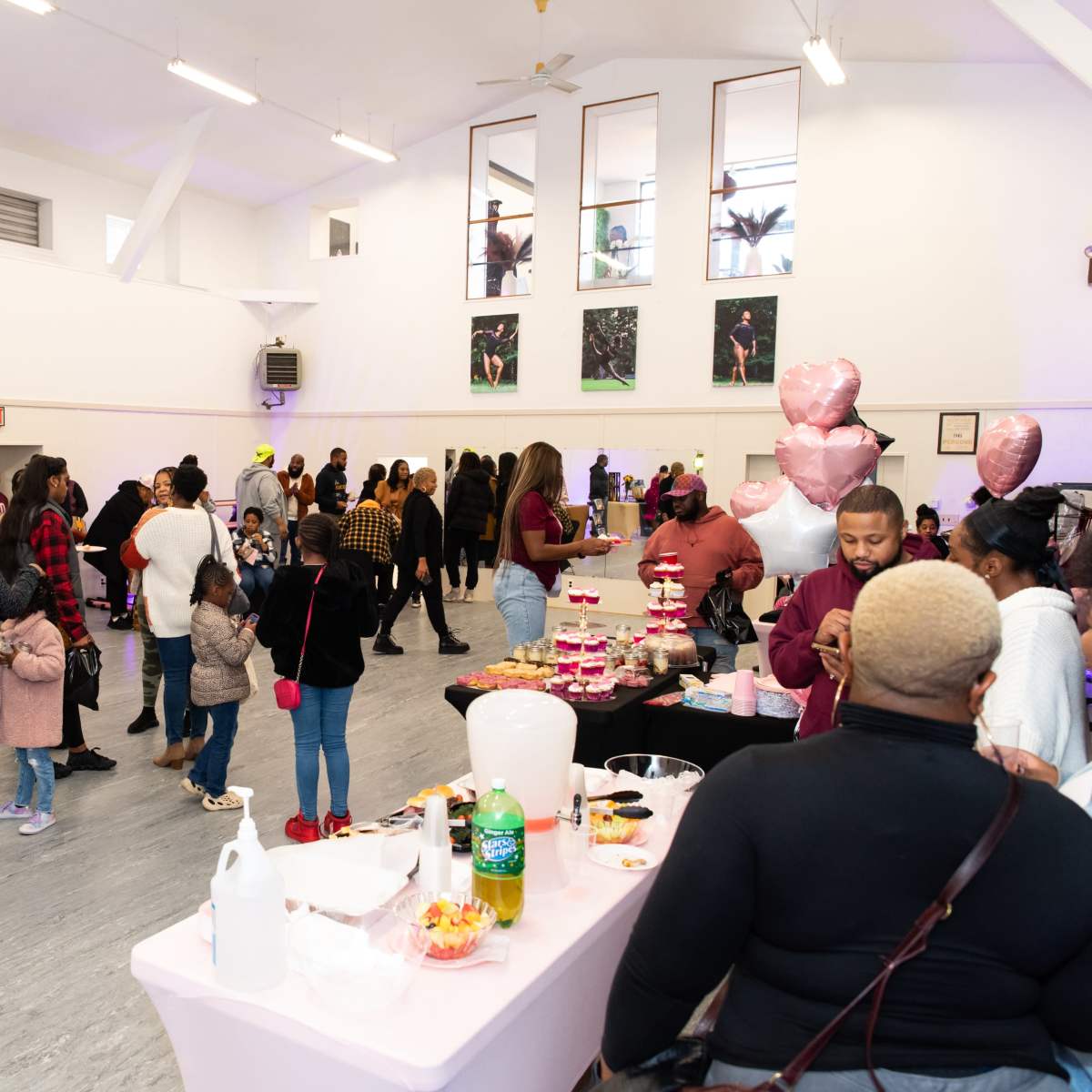 A large indoor gathering features various tables with food and decorations, while attendees socialize and interact throughout the spacious room.