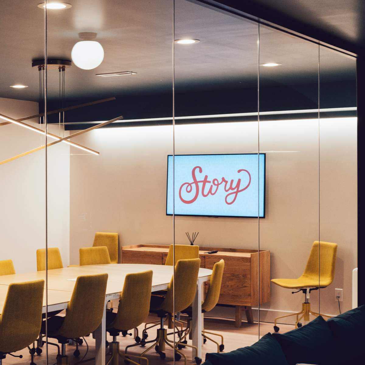 A modern conference room features a large table surrounded by yellow chairs, with a screen displaying the word "Story" on the wall.
