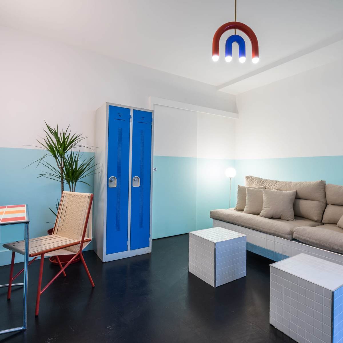 A modern room featuring a light blue and white color scheme, a beige sectional sofa, two white cube tables, a metal chair, and blue lockers.