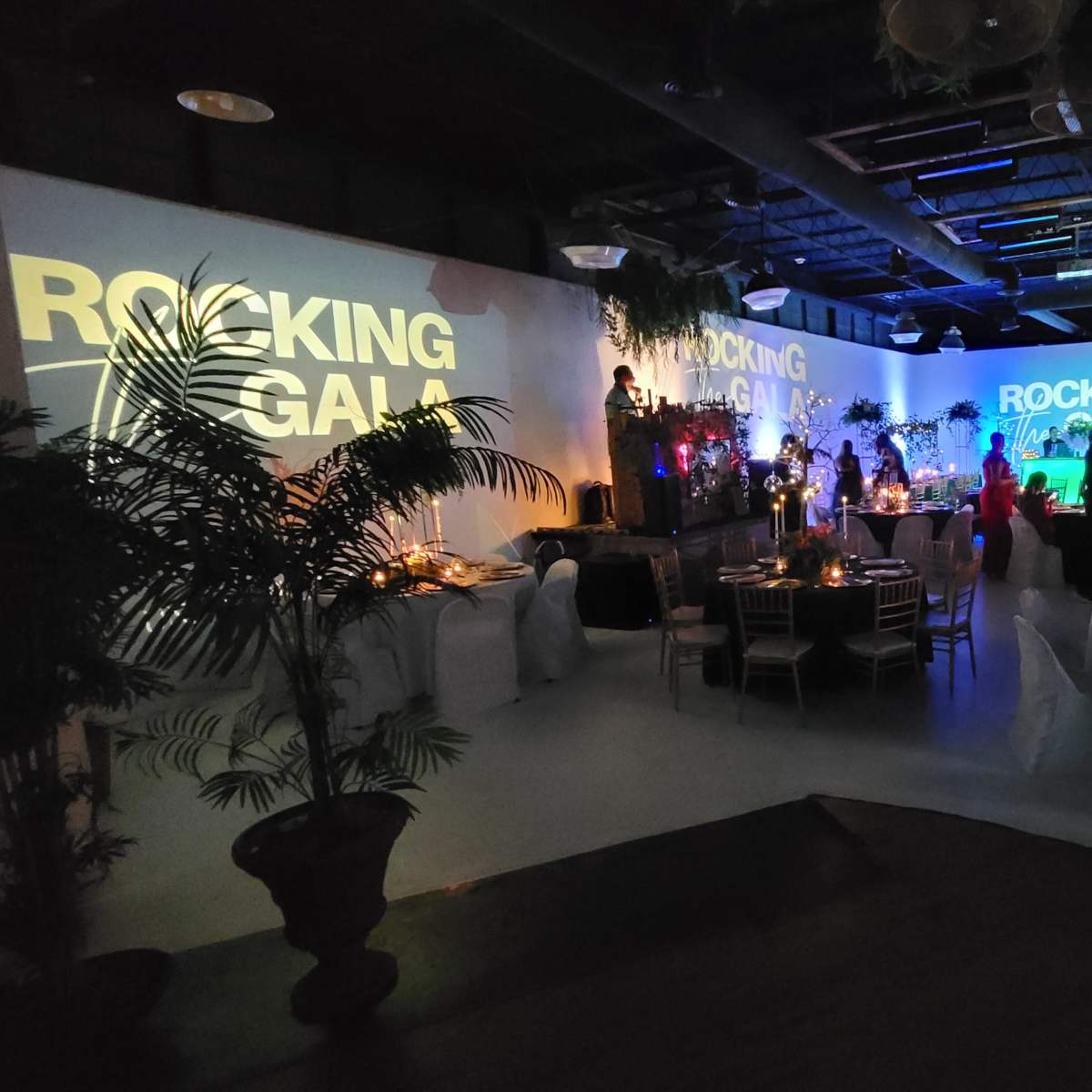 The indoor venue is set up for a gala, featuring elegantly arranged tables with candles, tropical plants, and a large projection displaying "ROCKING GALA" on the walls.
