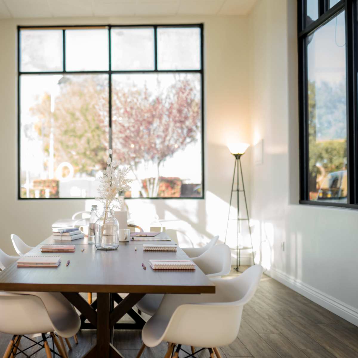 A spacious conference room features a long wooden table surrounded by white chairs and large windows allowing natural light to enter.