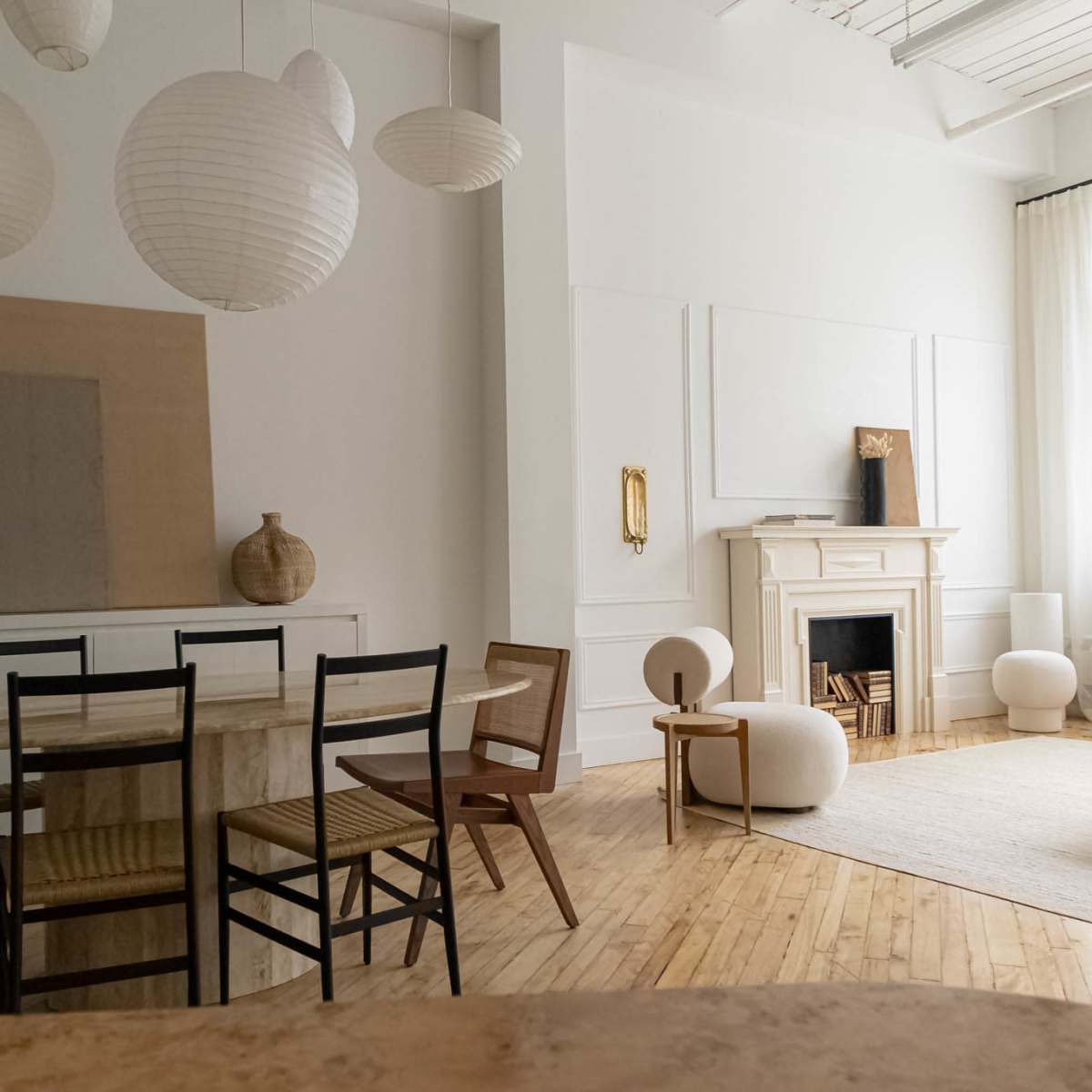 A spacious, minimalist living area features a dining table with chairs, a soft-colored rug, and a fireplace, illuminated by natural light through large windows.
