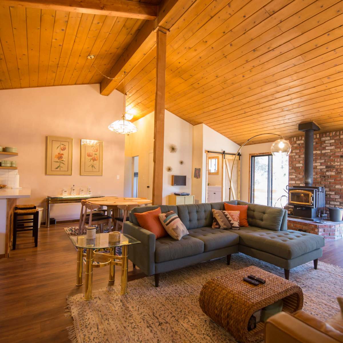 A modern living room features a sectional sofa, a dining area, and a wood-burning stove with large windows overlooking a forested view.
