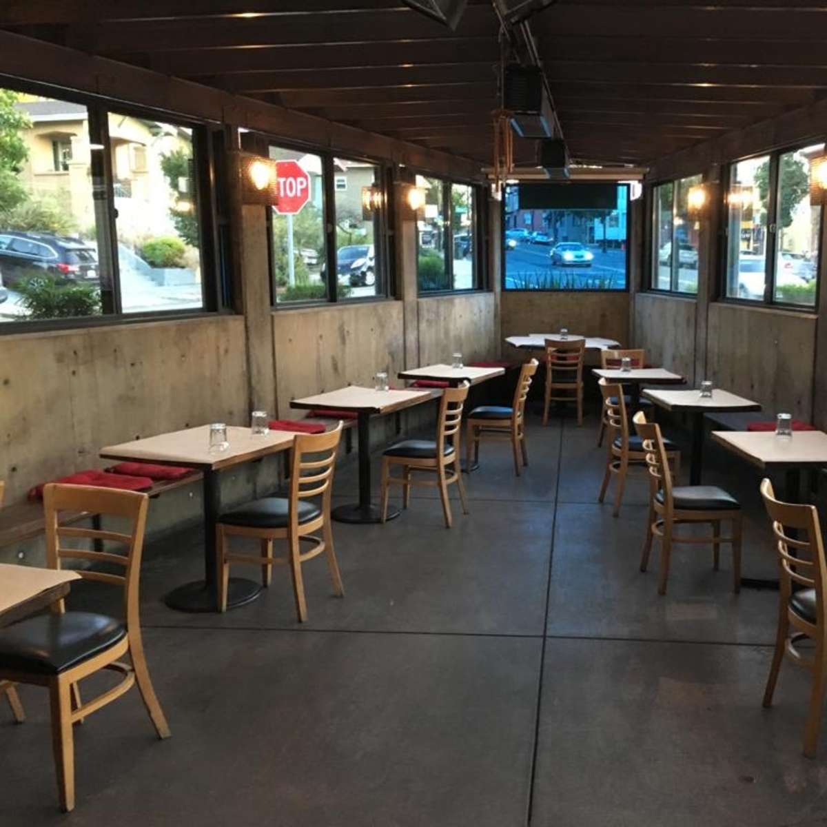A restaurant interior with several empty tables and chairs arranged in a long, narrow space enclosed by glass windows.