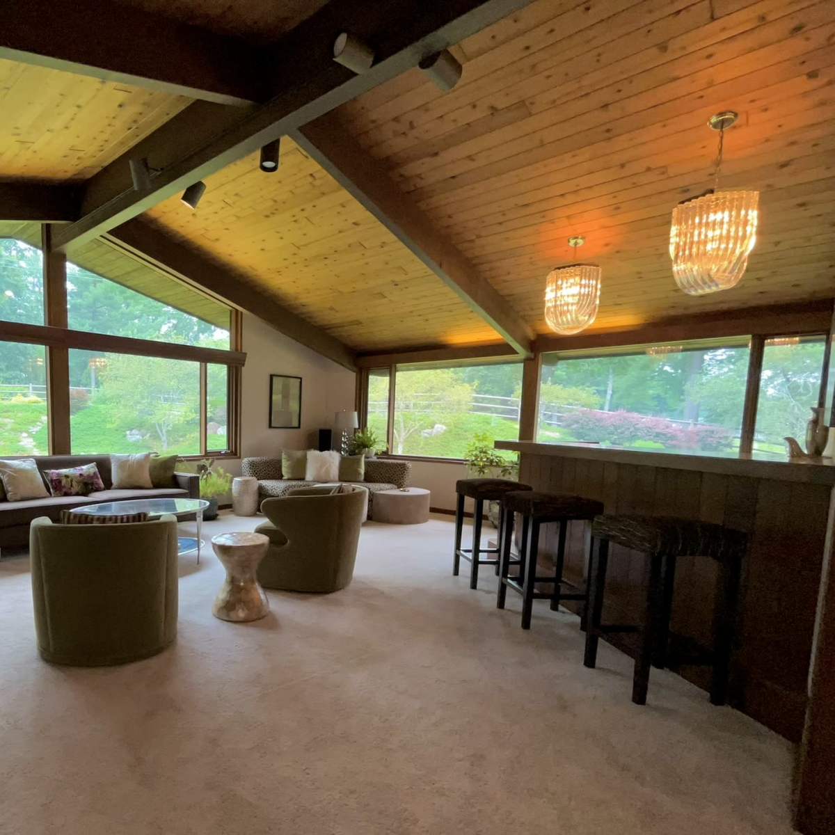 A spacious living room with large windows, wooden beams, and a bar area featuring stools.