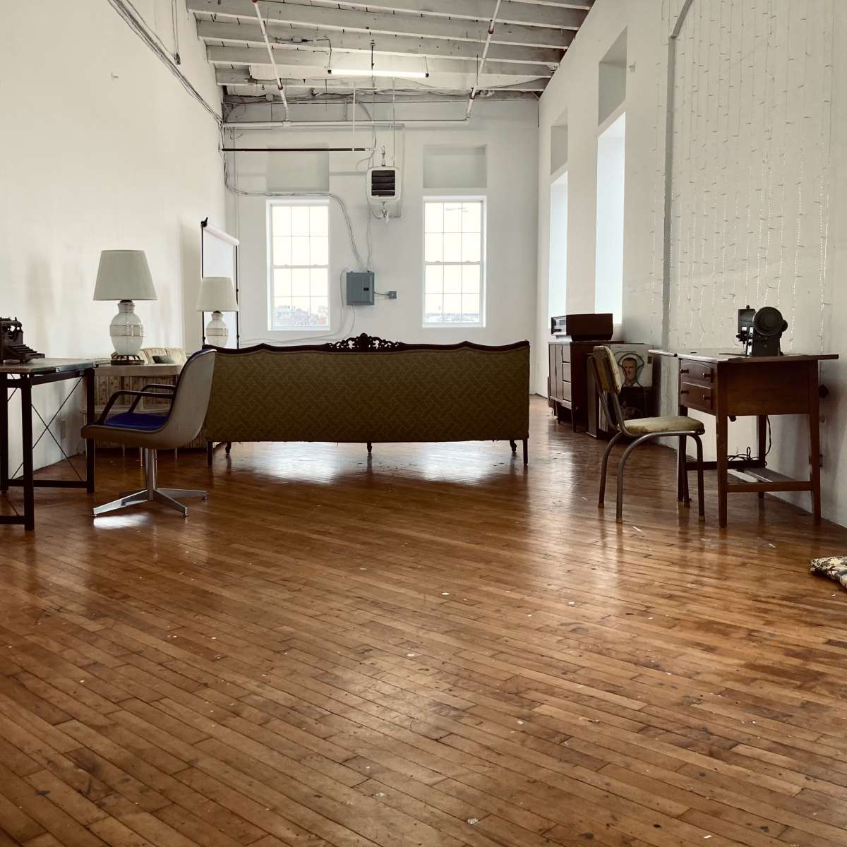 A spacious, minimally furnished room with wooden floors, featuring a green couch, a lamp, a chair, and a couple of tables.
