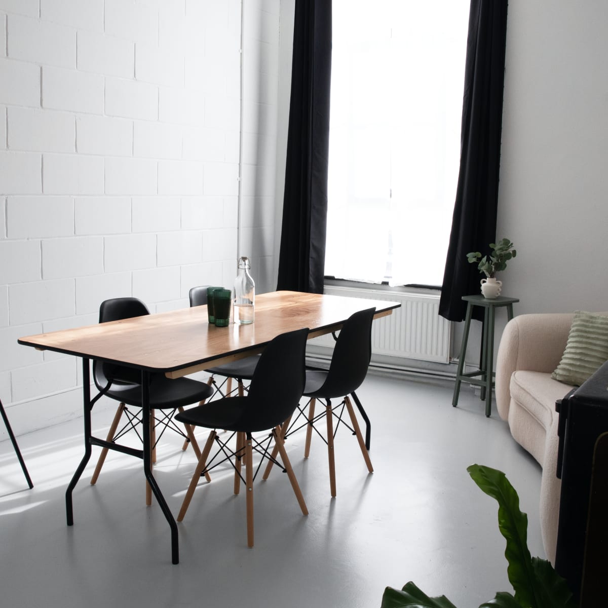 A bright, minimalist meeting room features a wooden table surrounded by black chairs, a whiteboard on an easel, and a sofa against the wall.